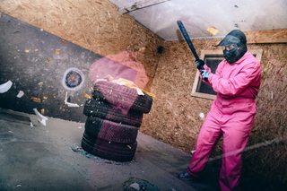 Eine Frau in einem pinken Overall und mit schwarzem Baseballschläger in der Hand im Rage-Room.